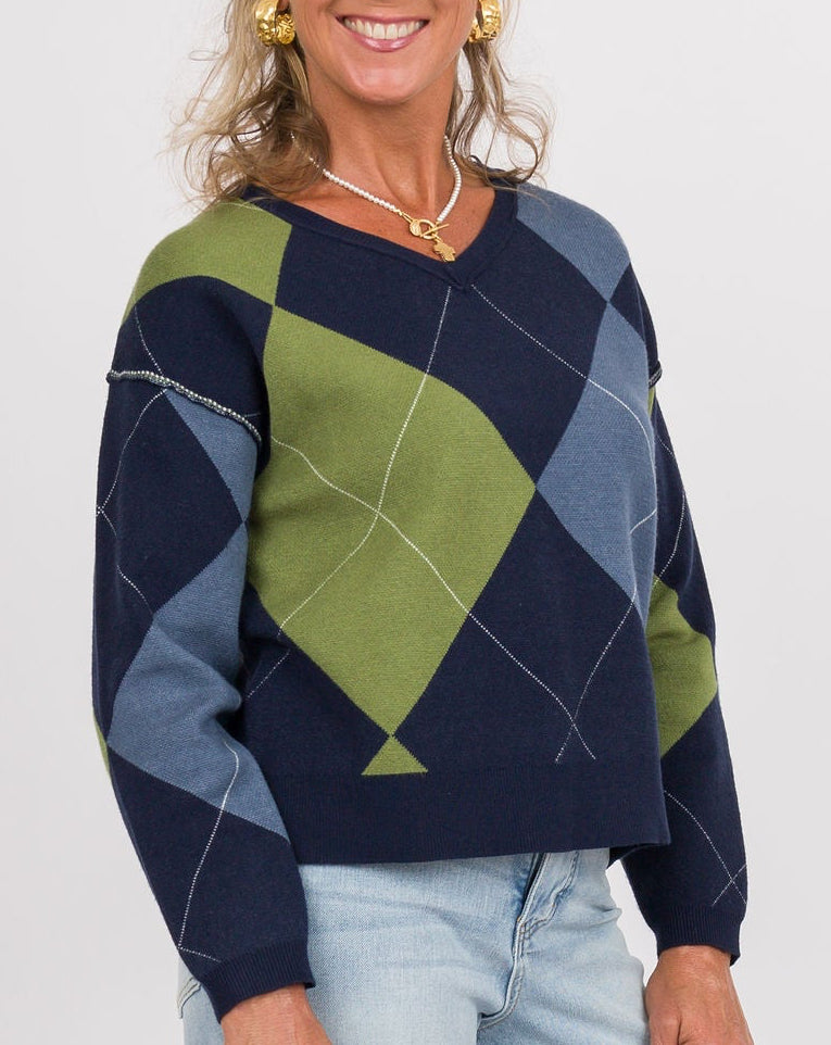 A smiling woman stands against a light gray studio background wearing a navy, green, and blue argyle sweater, light-wash wide-leg jeans, tan shoes, and holding a beige clutch.