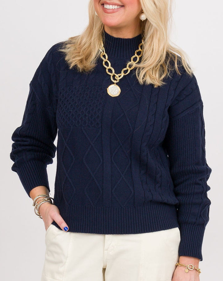A woman modeling a navy blue cable-knit mock-neck sweater, cream wide-leg trousers, brown clogs, and holding a small plaid clutch against a light gray studio background.
