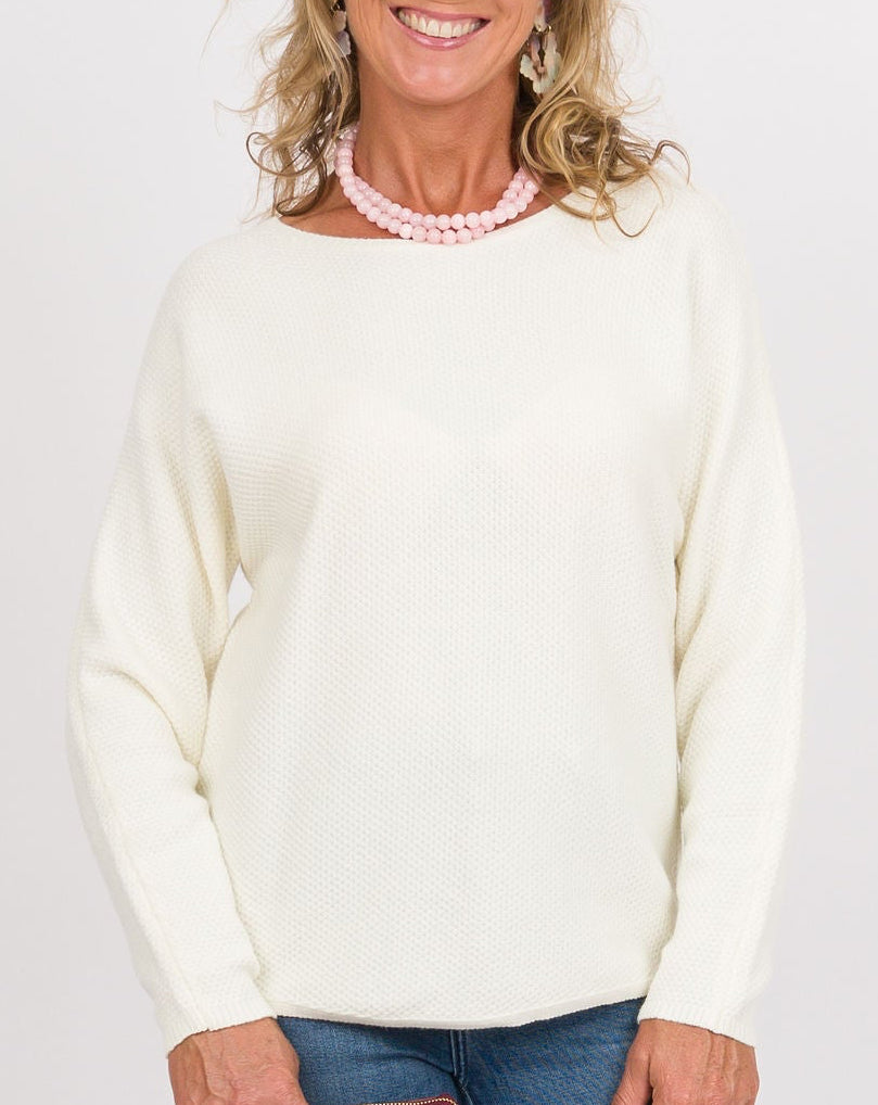 A woman modeling a cream knit sweater and blue flared jeans, holding a burgundy clutch against a plain light gray studio background.