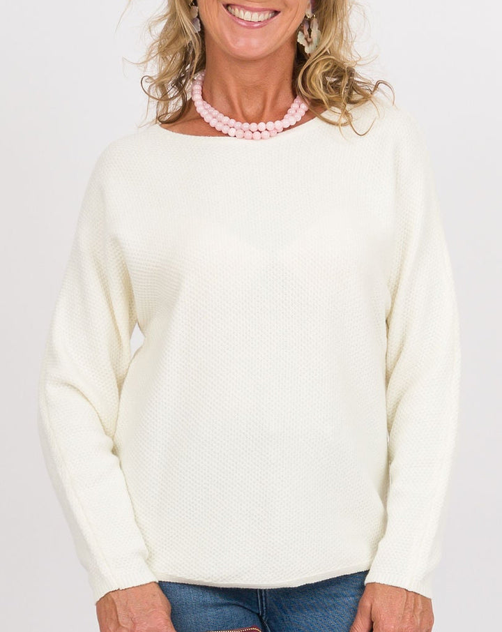 A woman modeling a cream knit sweater and blue flared jeans, holding a burgundy clutch against a plain light gray studio background.