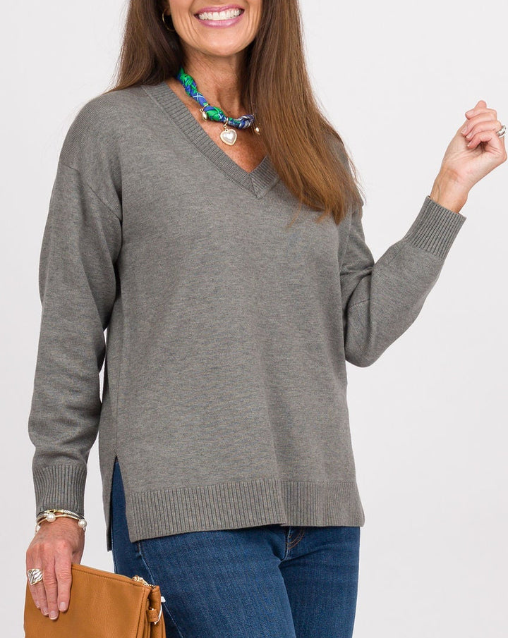 A woman modeling a gray long-sleeve knit sweater, blue jeans, tan clogs, and holding a tan clutch against a light gray studio background.