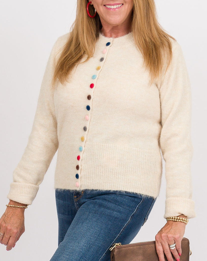 A woman modeling a cream knit cardigan with colorful button details, paired with blue wide-leg jeans, tan clogs, and holding a brown leather clutch against a plain white studio background.