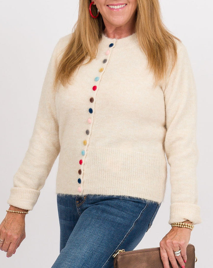 A woman modeling a cream knit cardigan with colorful button details, paired with blue wide-leg jeans, tan clogs, and holding a brown leather clutch against a plain white studio background.
