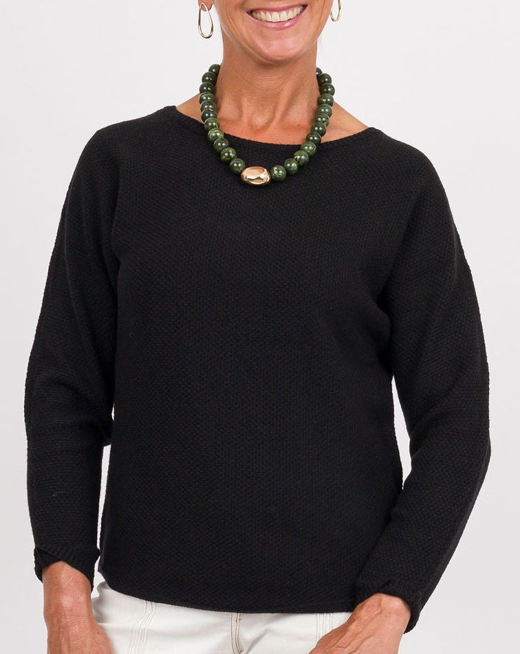 A smiling older woman stands against a light gray studio background wearing a black long-sleeve top, white wide-leg pants, dark flats, a green beaded necklace, and holding a rust-brown clutch.