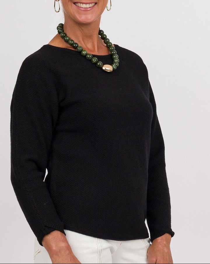 A smiling older woman stands against a light gray studio background wearing a black long-sleeve top, wide-leg white pants, brown shoes, a green beaded necklace, and holding a brown leather clutch.