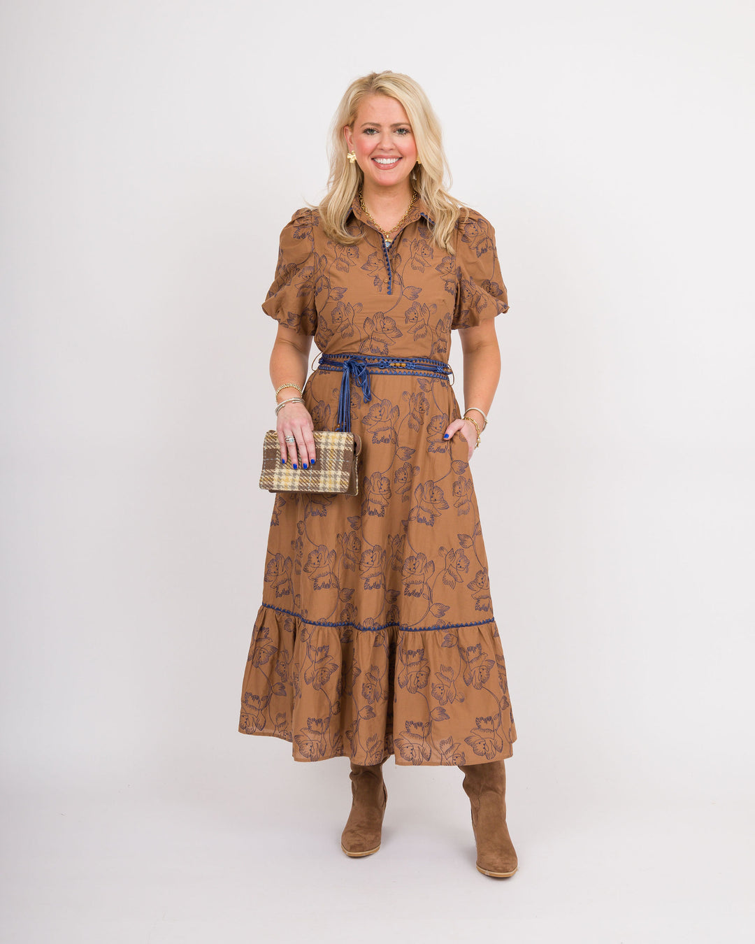 A woman modeling a brown midi dress with a blue patterned print, a blue tie belt, and brown suede boots, holding a plaid clutch against a plain white studio background.