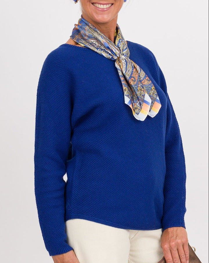A woman stands against a light gray studio background wearing a bright blue sweater, cream wide-leg pants, a multicolor patterned scarf, and brown shoes, while holding a brown clutch.