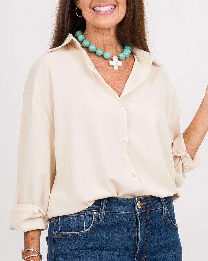 A woman modeling a cream long-sleeve button-up shirt, blue flared jeans, tan pointed-toe flats, a turquoise necklace, and carrying a brown leather handbag against a light gray studio background.