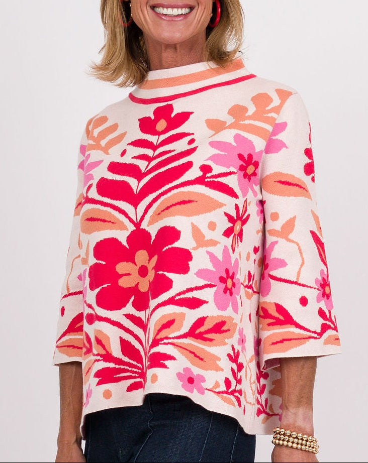 A woman modeling a vibrant floral print knit top with three-quarter sleeves, paired with dark blue wide-leg jeans, tan suede boots, and holding a brown clutch against a plain white studio background.
