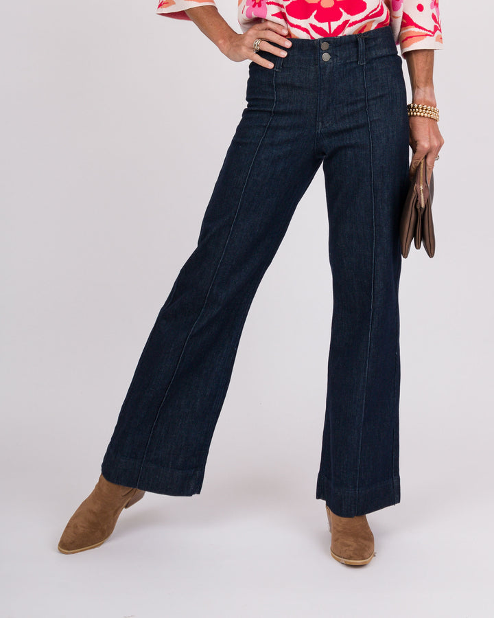 A person modeling dark blue wide-leg denim jeans with a floral pink and white top, brown suede boots, and holding a brown clutch against a light gray studio background.