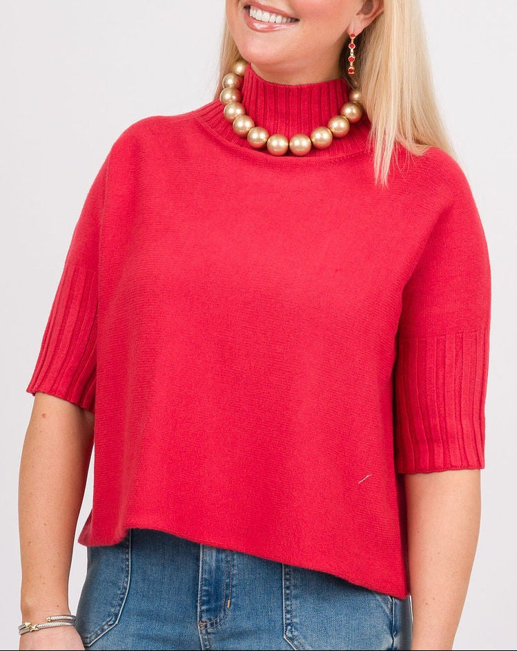 A woman modeling a bright red short-sleeve mock neck knit top, paired with blue jeans, a brown woven shoulder bag, and brown shoes, against a light gray studio background.