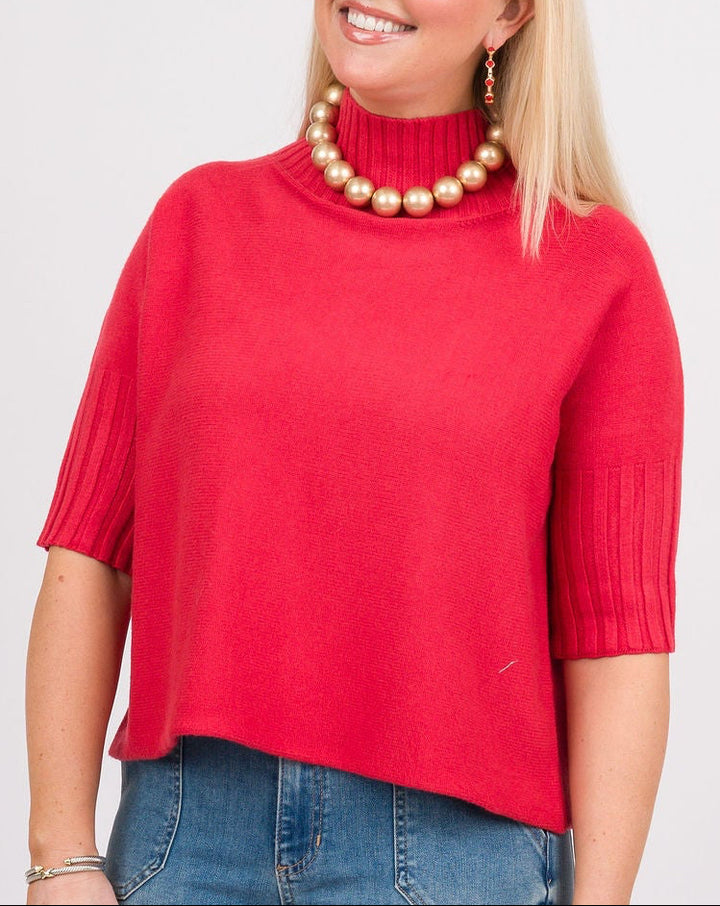 A woman modeling a bright red short-sleeve mock neck knit top, paired with blue jeans, a brown woven shoulder bag, and brown shoes, against a light gray studio background.