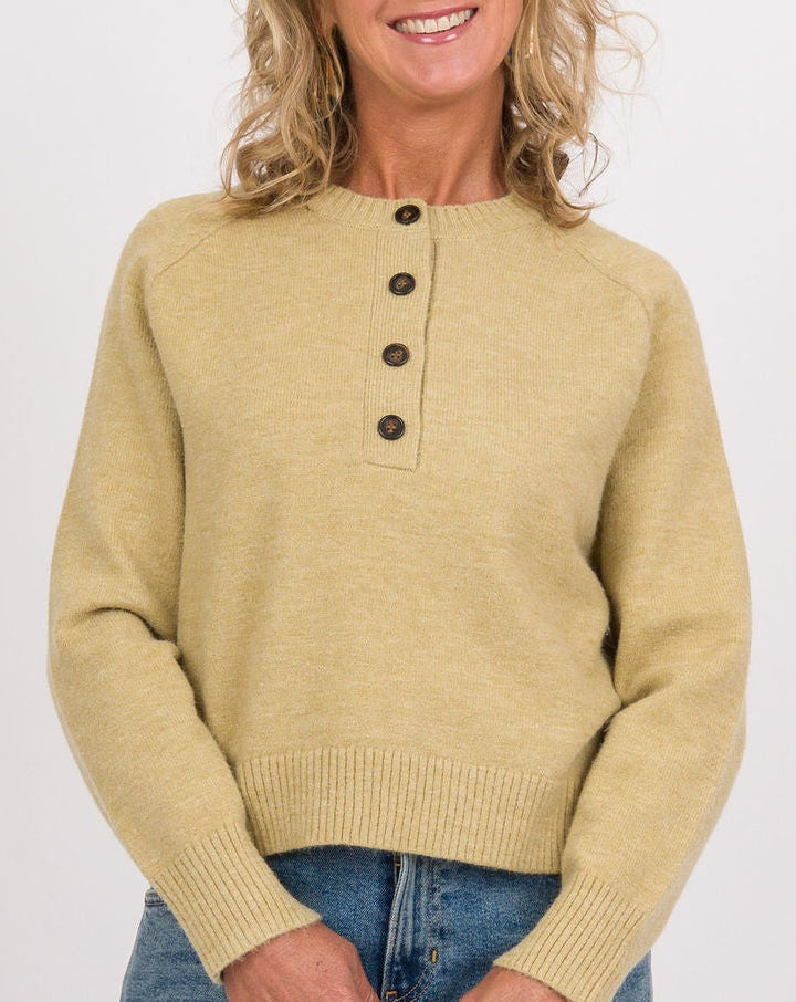 A woman stands against a light green studio background wearing a pale yellow knit henley sweater, wide-leg blue jeans, navy sneakers, and holding a small taupe and cream handbag.
