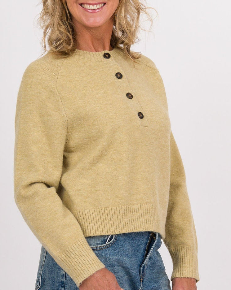 A woman modeling a light green cropped knit sweater with dark buttons, blue jeans, and navy sneakers, holding a beige and white handbag against a plain light gray studio background.
