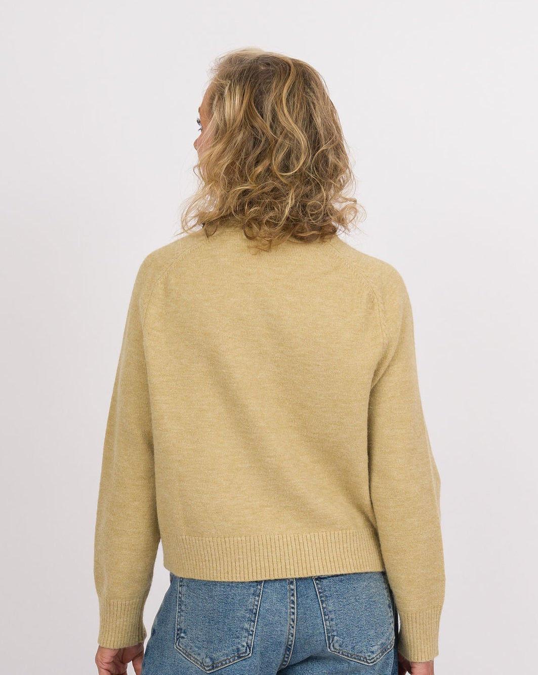 A model shown from the back wearing a light yellow knit sweater, wide-leg blue denim jeans, and black slip-on shoes with beige soles, set against a plain white studio background.