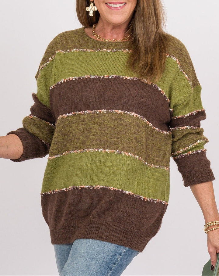 A woman modeling a green and brown striped knit sweater, blue flared jeans, and brown suede boots, holding a green clutch against a plain light gray studio background.