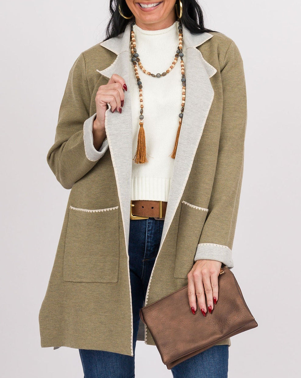 A woman modeling a mid-length olive green coat with cream trim over a cream knit top, blue jeans, tan boots, and holding a brown clutch against a light gray studio background.