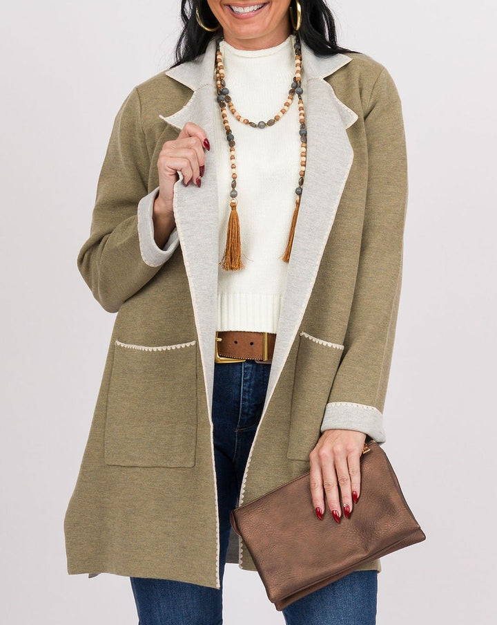 A woman modeling a mid-length olive green coat with cream trim over a cream knit top, blue jeans, tan boots, and holding a brown clutch against a light gray studio background.