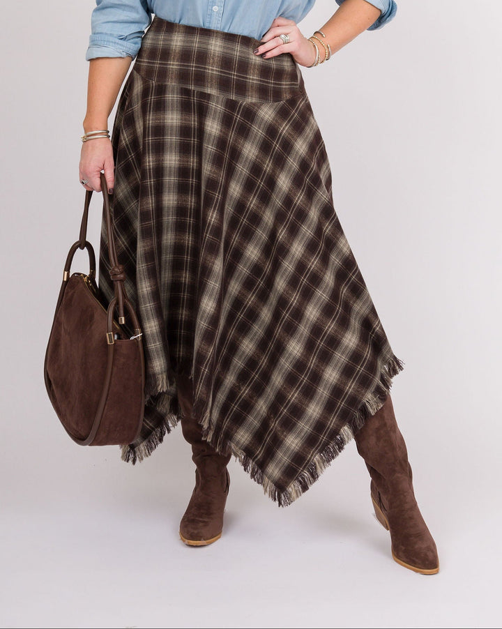 A woman modeling a brown and beige plaid wrap skirt with fringe, styled with a light blue button-down shirt, brown suede boots, and a brown handbag against a light gray studio background.