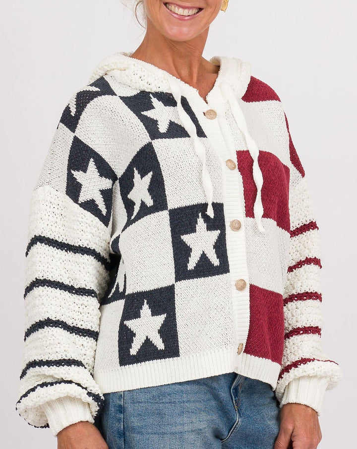 A smiling woman stands against a light gray studio background wearing a knit cardigan with a black, white, and burgundy American flag star pattern, blue jeans, navy sneakers, and carrying a brown leather handbag.