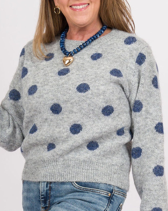 A woman modeling a light gray knit sweater with navy blue polka dots, paired with blue jeans, brown clogs, and holding a blue woven handbag with a gold chain strap against a plain white studio background.