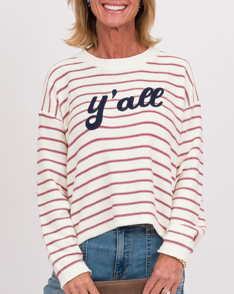 A woman modeling a cream and red striped long-sleeve crewneck sweater with 'y'all' written across the chest in navy blue, paired with blue jeans and holding a brown clutch against a plain white studio background.
