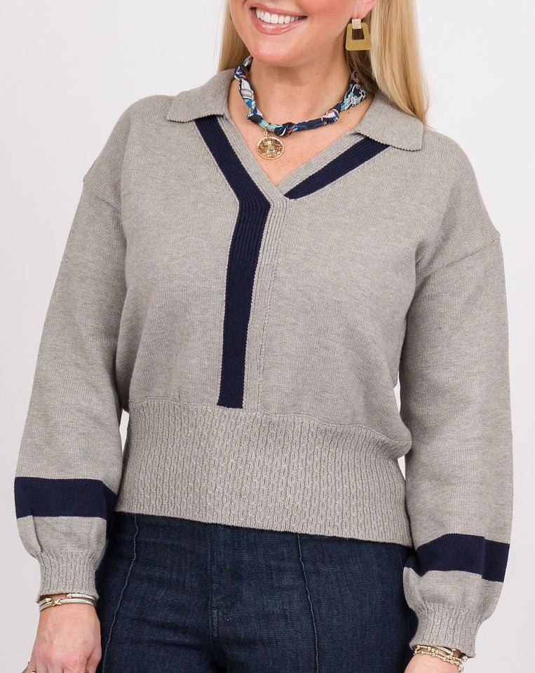 A woman modeling a light gray knit sweater with navy blue trim and dark blue wide-leg jeans, holding a beige clutch against a plain white studio background.