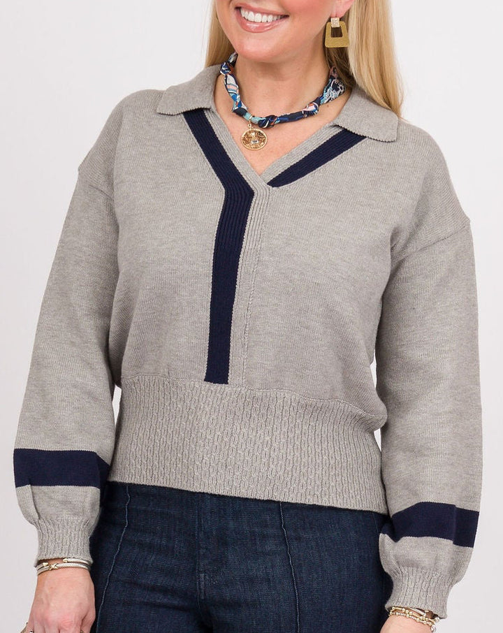 A woman modeling a light gray knit sweater with navy blue trim and dark blue wide-leg jeans, holding a beige clutch against a plain white studio background.