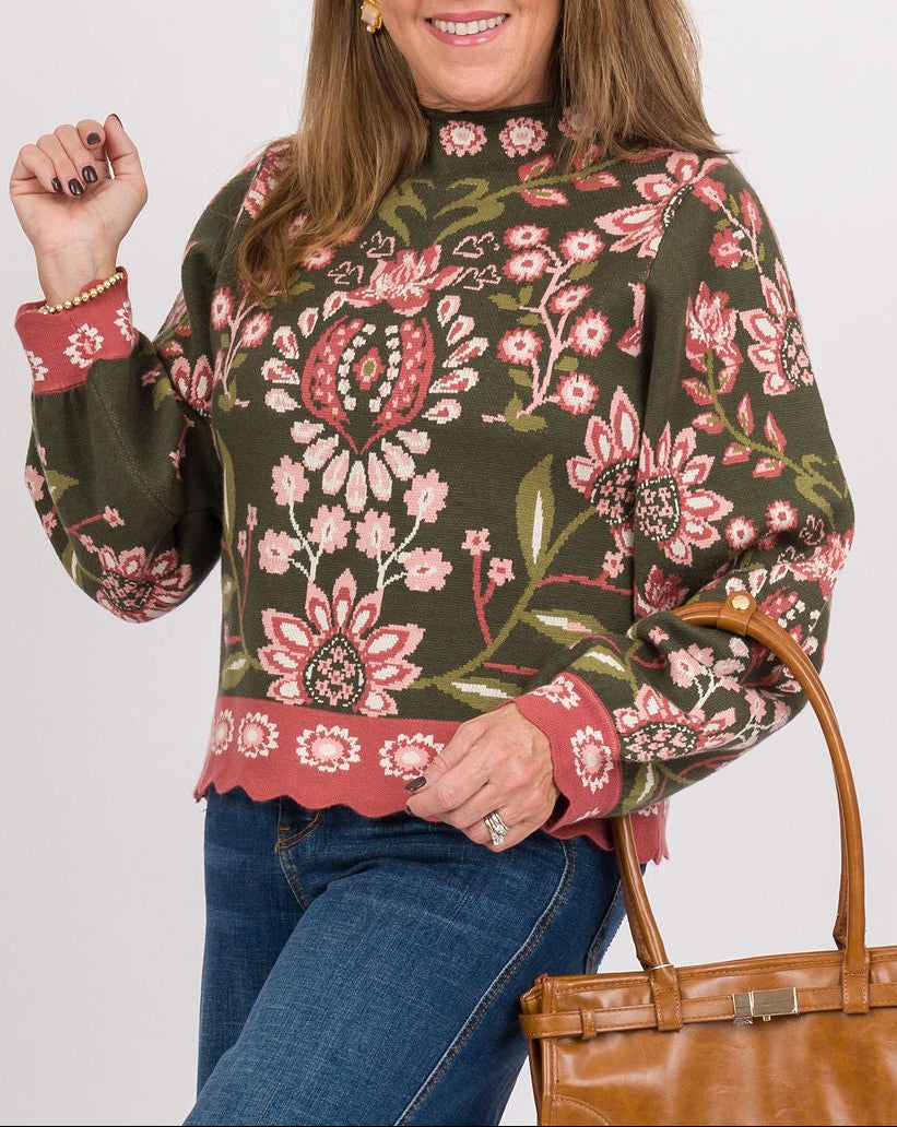 A woman modeling a green floral long-sleeve sweater, blue wide-leg jeans, tan clogs, and holding a brown leather handbag against a light gray studio background.