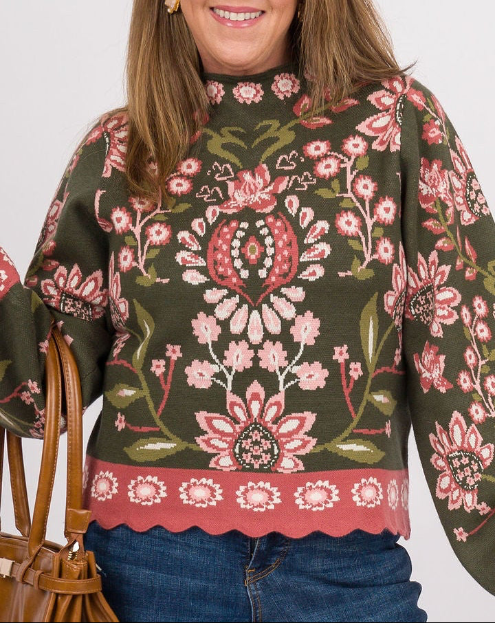 A woman modeling a dark green long-sleeve sweater with a pink and white floral pattern, paired with blue wide-leg jeans, brown clogs, and a brown leather handbag against a light gray studio background.
