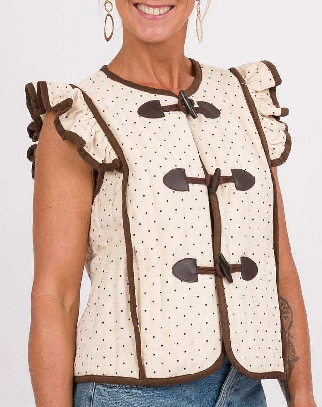 A woman modeling a sleeveless cream vest with brown trim and toggle closures, paired with blue jeans, brown shoes, and holding a brown shoulder bag against a light gray studio background.