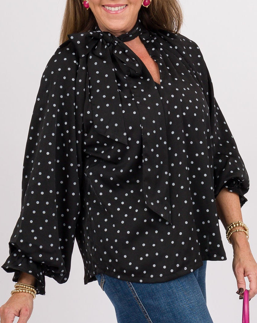 A woman modeling a black long-sleeve blouse with white polka dots, paired with blue wide-leg jeans, black ankle boots, and holding a pink clutch bag against a plain light gray studio background.