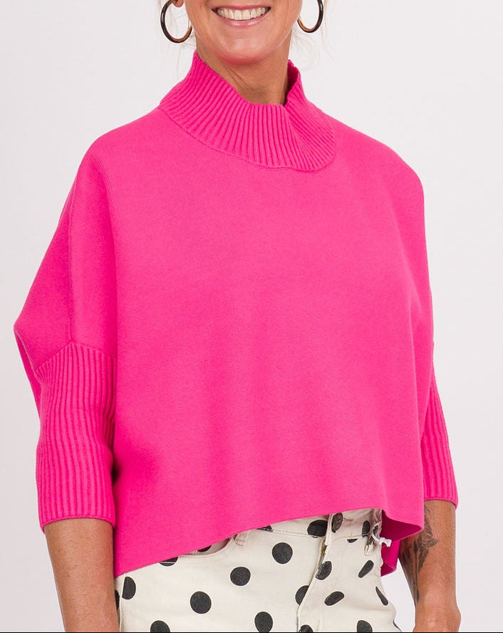 A woman modeling a bright pink mock-neck knit sweater, white pants with black polka dots, and pink sneakers, holding a beige clutch bag against a plain white studio background.
