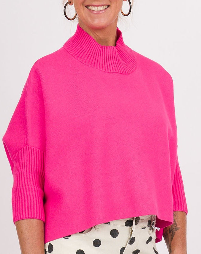 A woman modeling a bright pink mock-neck knit sweater, white pants with black polka dots, and pink sneakers, holding a beige clutch bag against a plain white studio background.