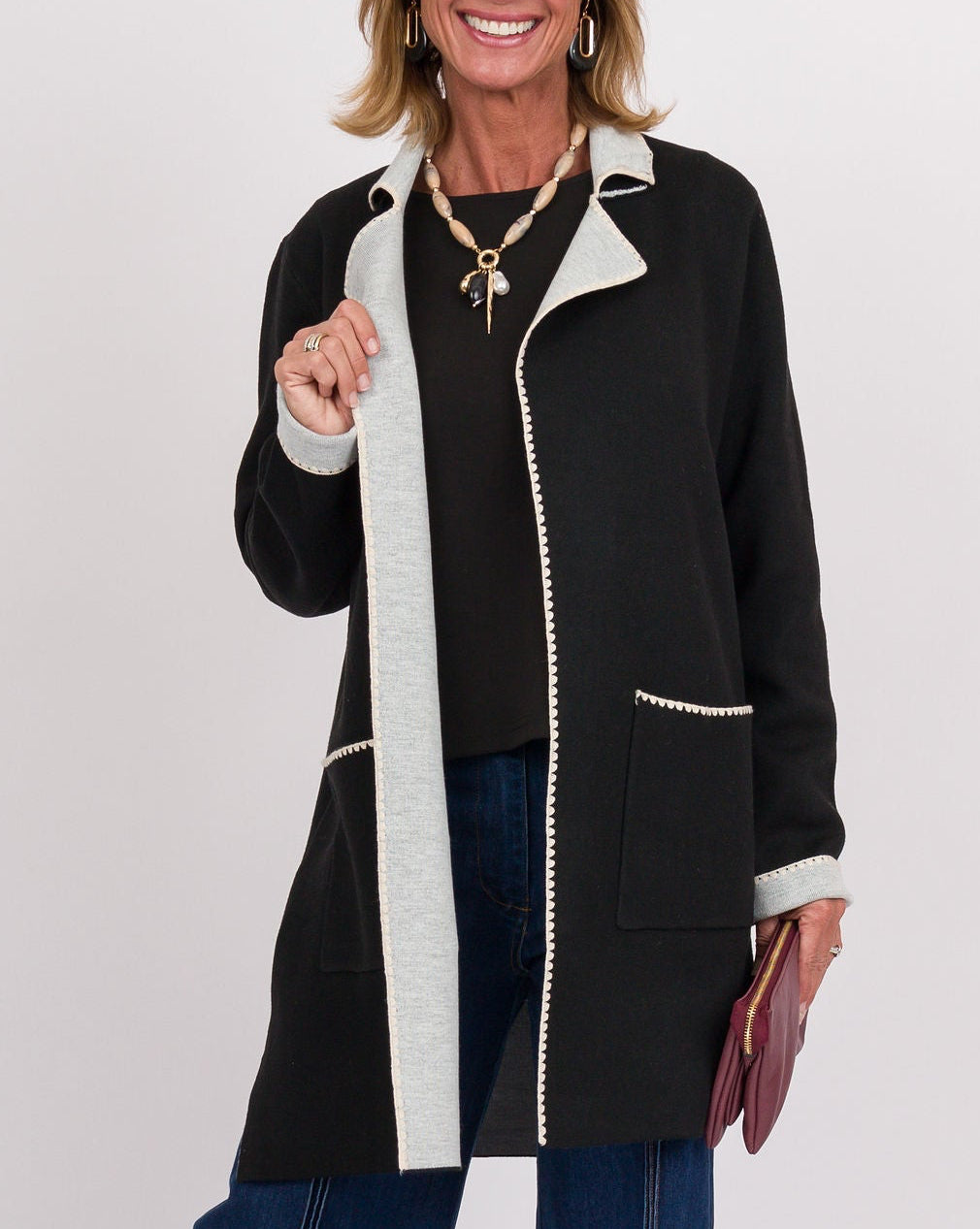 A smiling woman stands against a light gray studio background wearing a black mid-length zip-front coat with contrasting white trim and pockets, blue wide-leg jeans, and black shoes.