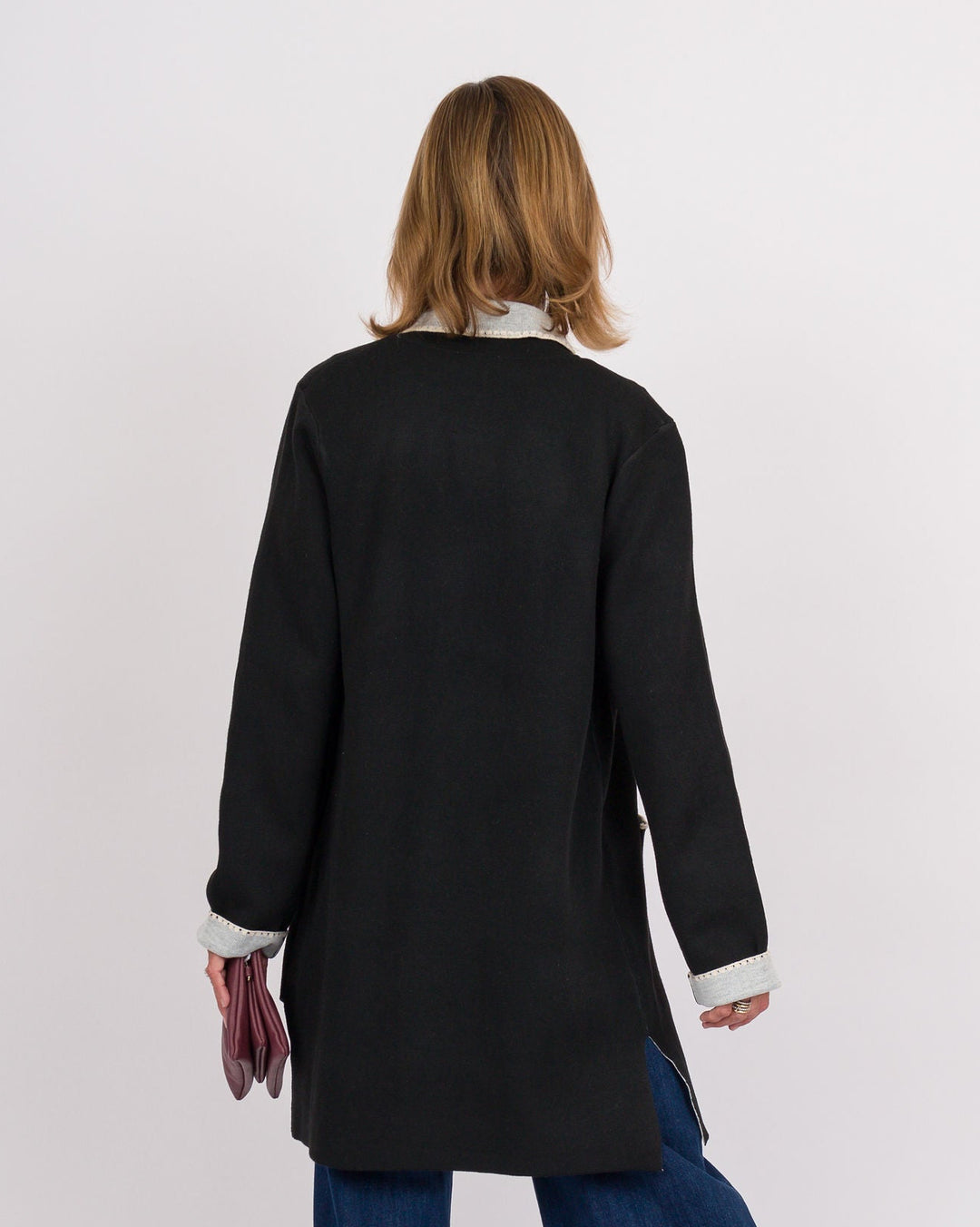 A woman is shown from behind wearing a black long-sleeve tunic-style cardigan over a white collared shirt, paired with wide-leg blue jeans, against a plain white studio background.