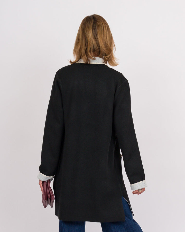 A woman is shown from behind wearing a black long-sleeve tunic-style cardigan over a white collared shirt, paired with wide-leg blue jeans, against a plain white studio background.