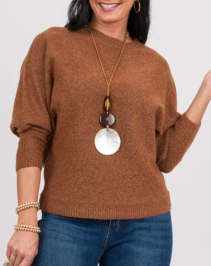 A woman modeling a brown knit sweater and blue jeans against a light gray studio background, holding a small plaid clutch.
