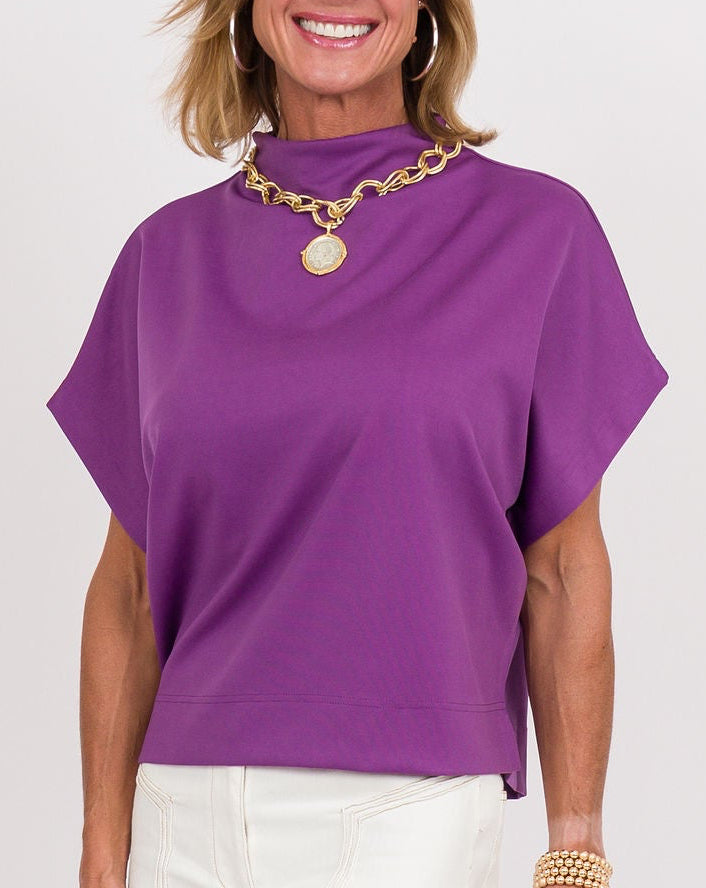 A woman modeling a purple short-sleeve mock neck top with white wide-leg pants, gold jewelry, and a brown clutch, standing against a plain white studio background.