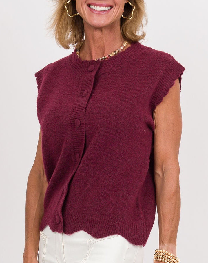 A woman modeling a sleeveless burgundy top and white wide-leg pants, holding a brown clutch against a plain white studio background.