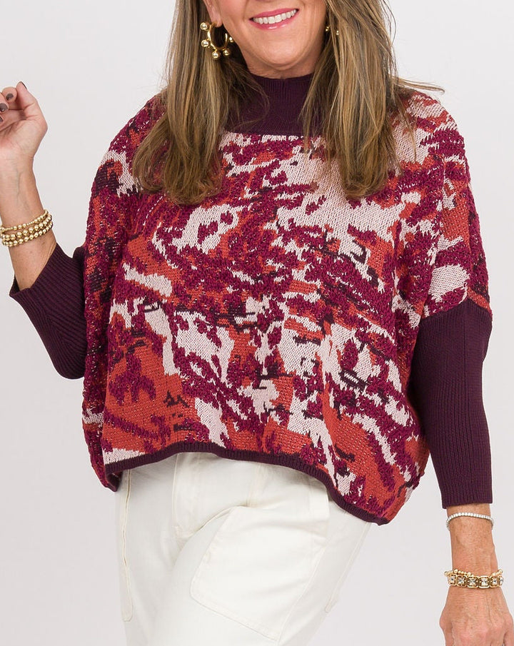 A woman modeling a long-sleeve knit top with a red, burgundy, and white abstract pattern over a dark burgundy base, paired with white wide-leg pants, clear block-heel sandals, and a beige clutch, against a plain white studio background.