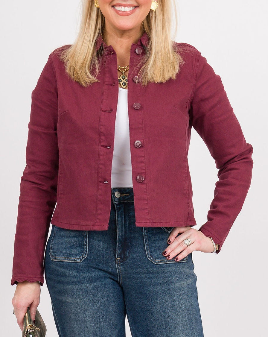 A woman modeling a cropped maroon button-up jacket, blue wide-leg jeans, and tan clogs, holding a small dark clutch against a light gray studio background.