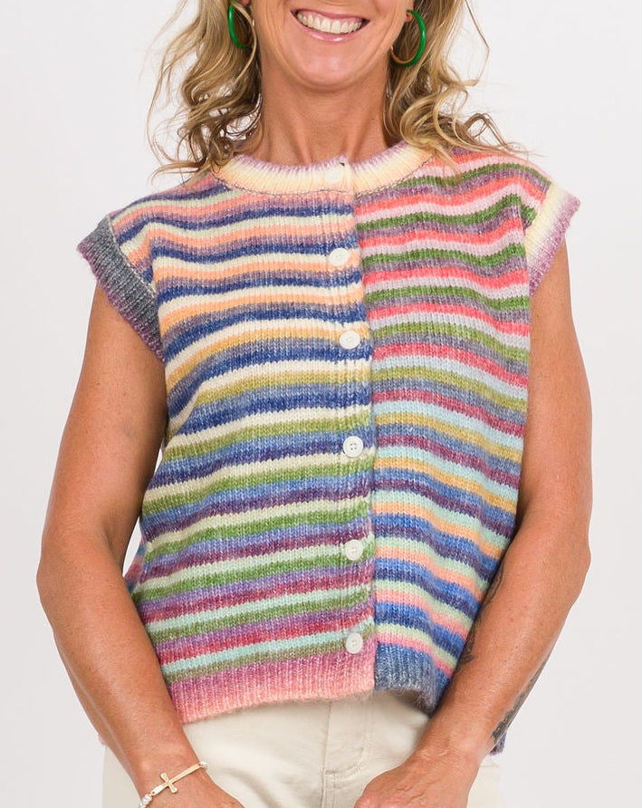 A woman stands against a plain white studio background wearing a colorful striped knit vest, white wide-leg pants, and clear high-heeled sandals, while holding a green clutch.