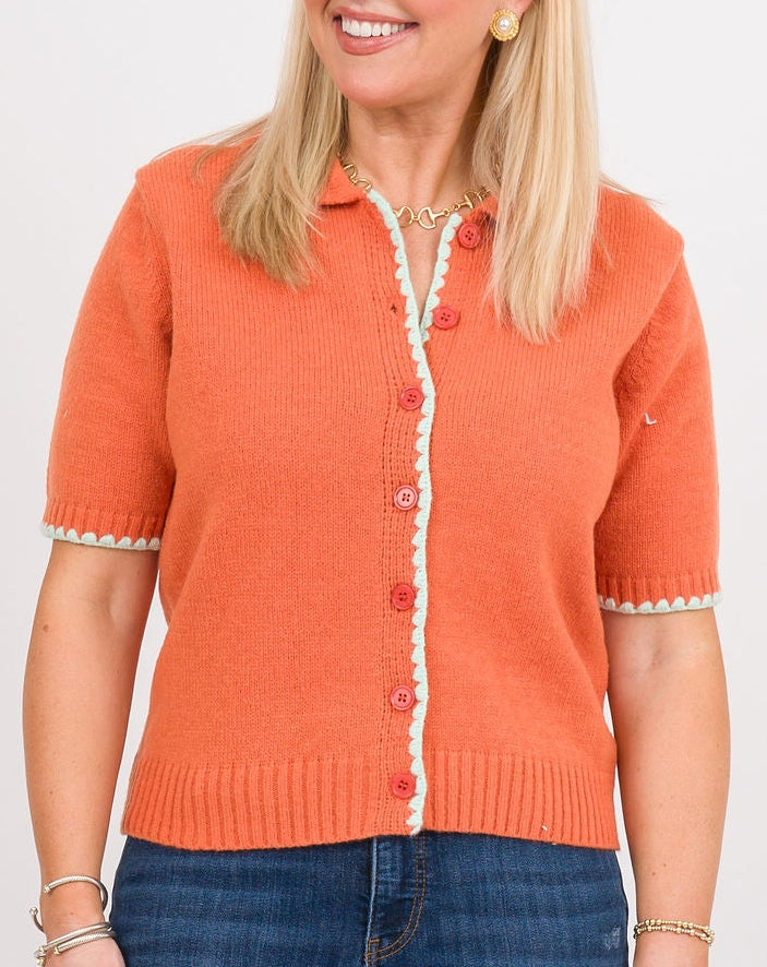 A woman modeling a short-sleeve orange knit polo cardigan with white trim and blue jeans, standing against a plain white studio background.