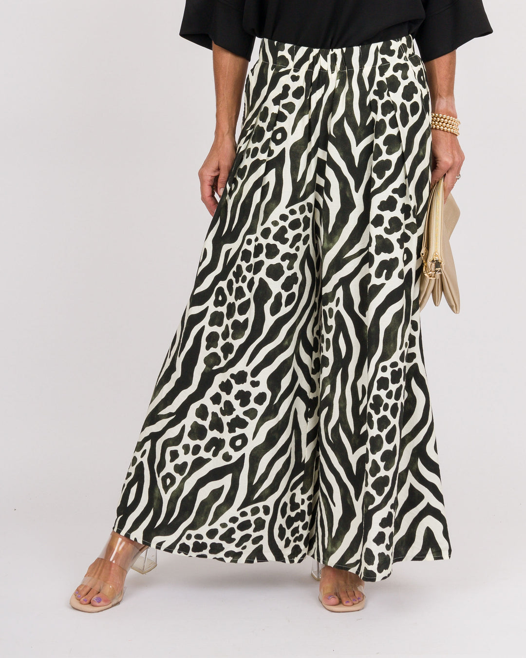 A person wearing wide-leg cropped pants with a bold black and off-white animal print, styled with a black top, clear-strap heels, and a beige clutch against a light gray studio background.
