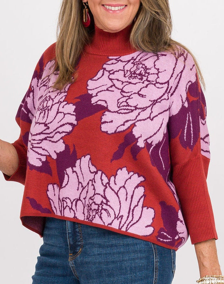 A woman modeling a red and purple floral print mock-neck top with blue wide-leg jeans, holding a beige clutch against a plain white studio background.