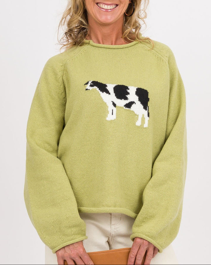 A smiling woman stands against a light gray studio background wearing a light olive green crewneck sweatshirt with a black and white cow graphic on the chest, paired with off-white wide-leg pants and tan flat shoes, while holding a tan clutch.