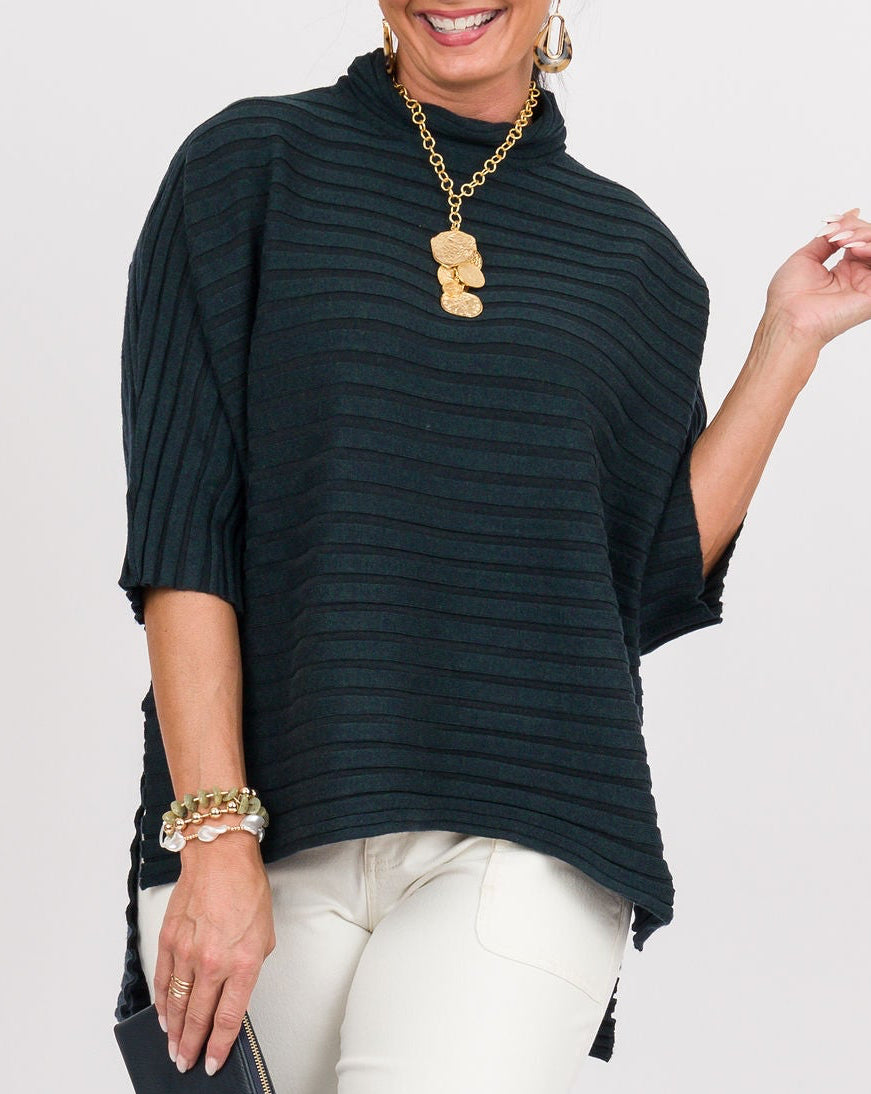 A woman modeling a dark green ribbed tunic top with elbow-length sleeves, paired with white cropped pants, gold jewelry, and tan sandals, standing against a light gray studio background.