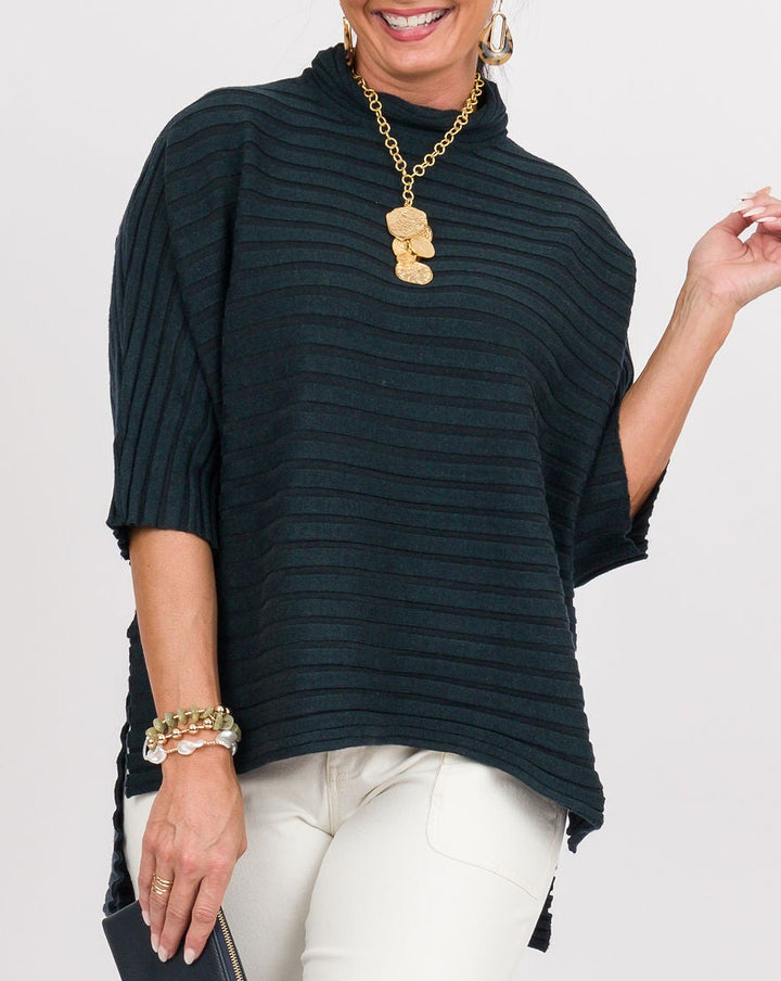 A woman modeling a dark green ribbed tunic top with elbow-length sleeves, paired with white cropped pants, gold jewelry, and tan sandals, standing against a light gray studio background.