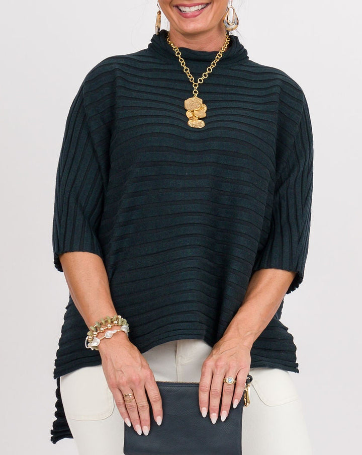 A woman modeling a dark green ribbed mock-neck top with tassel ties, paired with white cropped pants, clear slide sandals, and holding a small black clutch against a plain white studio background.